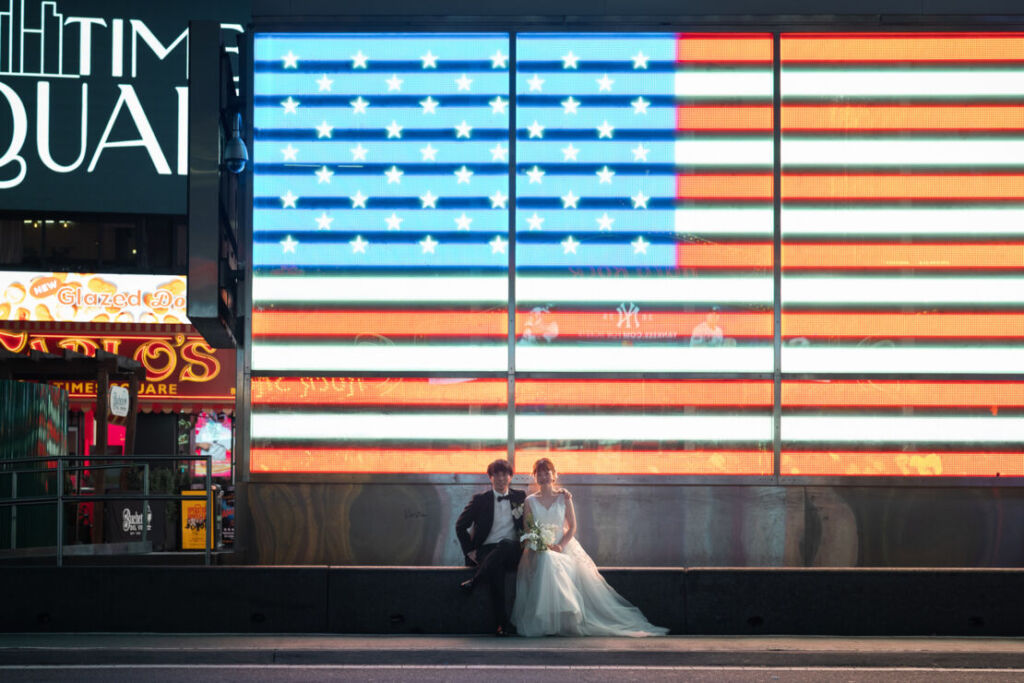 ニューヨークの前撮り|ウェディングフォト/タイムズスクエアの星条旗のネオンサインの前に座る花嫁と花婿。