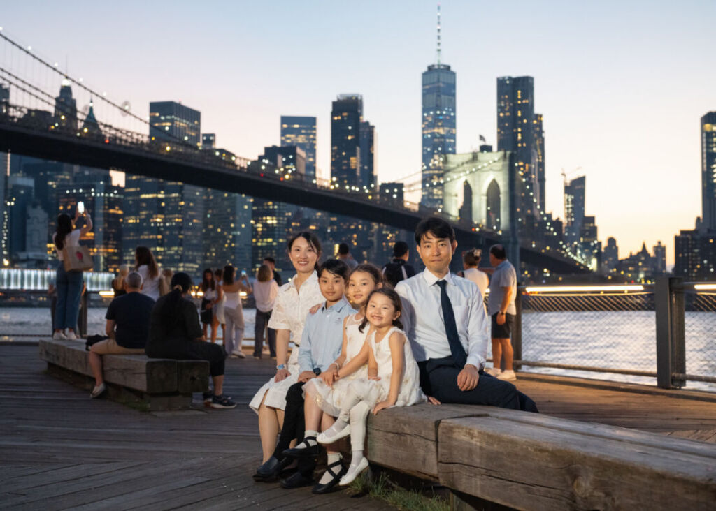 ニューヨークのファミリーフォト/グラデーションの空の向こうに夜景のマンハッタンが望めるブルックリンブリッジパークで。